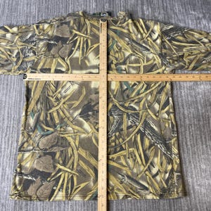 K&ouml;nnte beinhalten: Ein kurz&auml;rmeliges T-Shirt mit einem braunen, beigen und gr&uuml;nen Tarnmuster aus Bl&auml;ttern und Gras. Ein Holzlineal liegt vertikal und horizontal auf dem T-Shirt und zeigt eine Breite von 56 cm sowie eine L&auml;nge von 71 cm an.