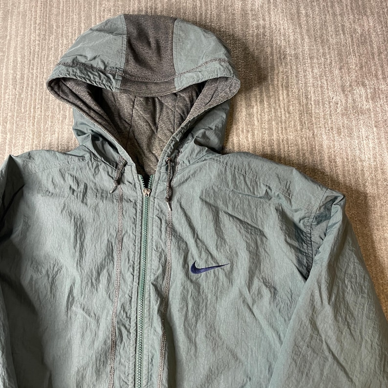 May include: A sage green zip-up hooded jacket featuring a dark gray quilted lining and hood panel. The jacket includes a full-length front zipper, a drawstring hood, and a small dark blue Nike swoosh logo embroidered on the left chest area.