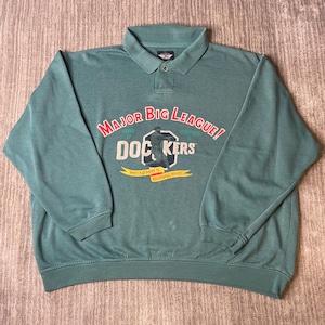May include: A sage green long-sleeve pullover shirt with a collar and a single button placket. The front features a graphic with the text "MAJOR BIG LEAGUE!", "DOCKERS", and "MY OLD MAN IS BASEBALL MAD!" alongside a baseball player illustration.