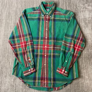 May include: A long-sleeve button-down shirt featuring a green, red, yellow, and navy blue plaid pattern. The shirt has a button-down collar, a single chest pocket, and white buttons. A GAP brand label is visible on the inside of the collar.