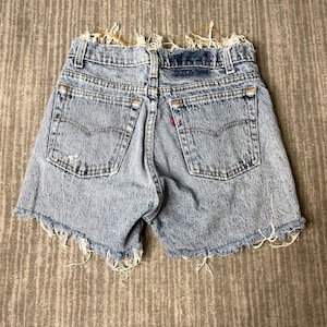 May include: Light blue denim shorts with frayed edges and back pockets. The shorts have a classic five-pocket design, a red Levi's tag, and a faded, worn appearance. The shorts are made of denim.
