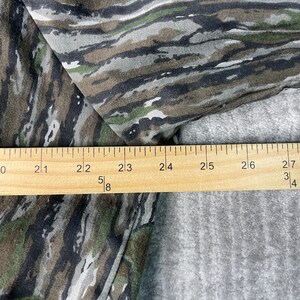May include: Camouflage pattern fabric in shades of green, brown, and gray. A wooden ruler is placed across the fabric, showing measurements in inches. The fabric has a textured appearance.