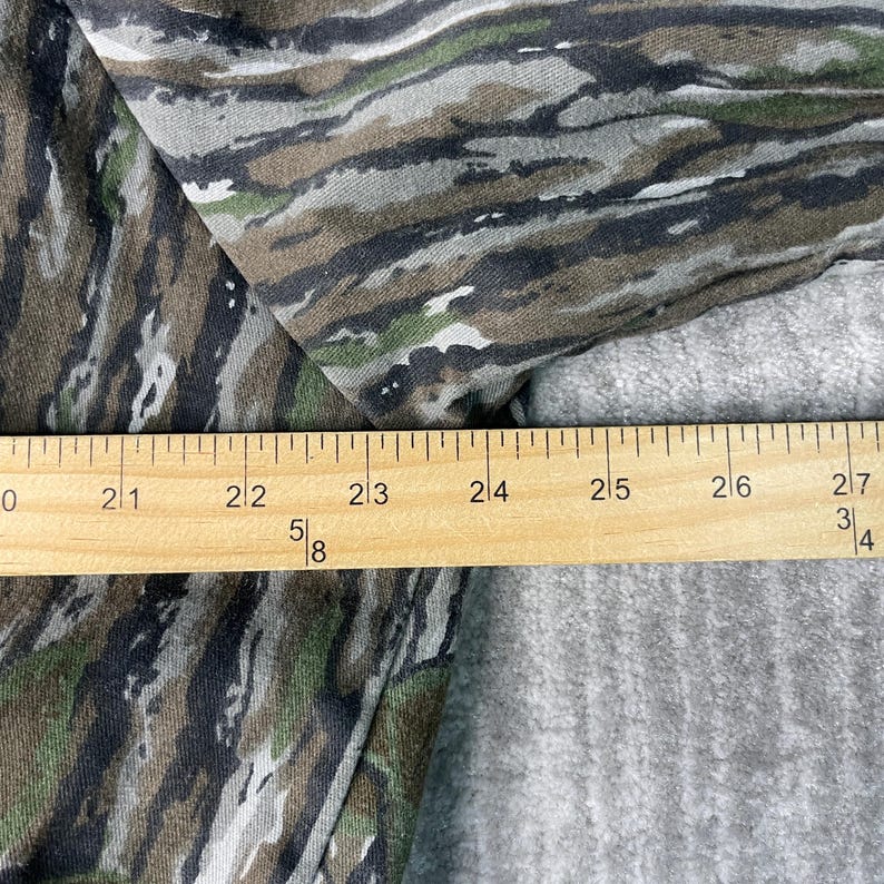 May include: Camouflage pattern fabric in shades of green, brown, and gray. A wooden ruler is placed across the fabric, showing measurements in inches. The fabric has a textured appearance.