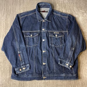 Pode incluir: Uma jaqueta jeans azul escuro com gola cl&aacute;ssica, fecho frontal de bot&otilde;es e dois bolsos no peito com abas abotoadas. A palavra AVIREX est&aacute; bordada em branco no bolso esquerdo e uma pequena etiqueta de marca est&aacute; na manga direita.