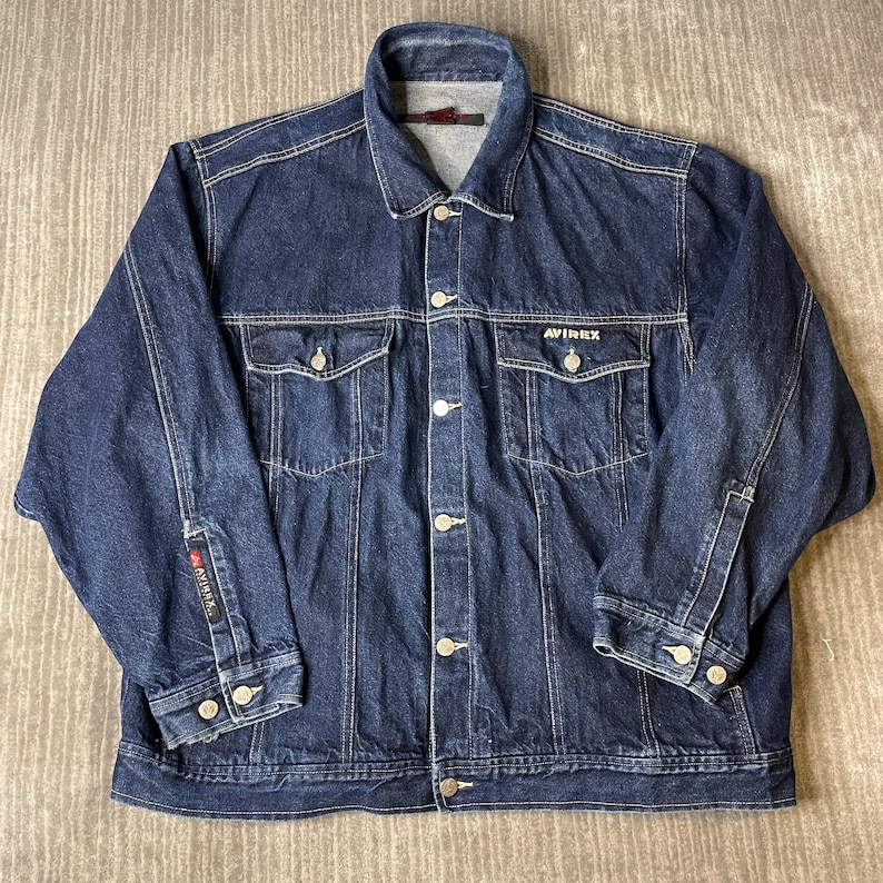 Pode incluir: Uma jaqueta jeans azul escuro com gola cl&aacute;ssica, fecho frontal de bot&otilde;es e dois bolsos no peito com abas abotoadas. A palavra AVIREX est&aacute; bordada em branco no bolso esquerdo e uma pequena etiqueta de marca est&aacute; na manga direita.