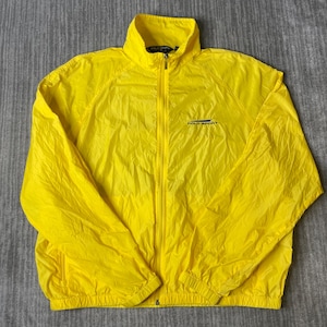May include: A bright yellow lightweight windbreaker jacket featuring a full-length front zipper, a high collar, and long sleeves with elastic cuffs. The text Polo Sport is printed in dark blue on the upper left chest area of the garment.