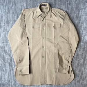 May include: A long-sleeved tan button-down shirt featuring a pointed collar, two chest pockets with buttoned flaps, and a full-length front button closure. The shirt includes buttoned cuffs and a curved hemline, with visible stitching details.
