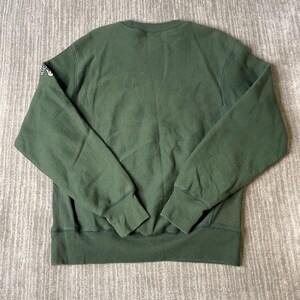 May include: A dark green sweatshirt with a crew neck and long sleeves. The cuffs and hem are ribbed. The left sleeve has a small white graphic. The sweatshirt is made of a soft, comfortable material.