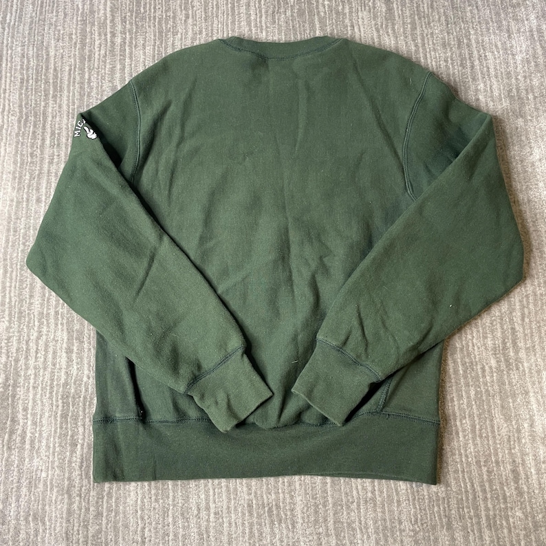 May include: A dark green sweatshirt with a crew neck and long sleeves. The cuffs and hem are ribbed. The left sleeve has a small white graphic. The sweatshirt is made of a soft, comfortable material.