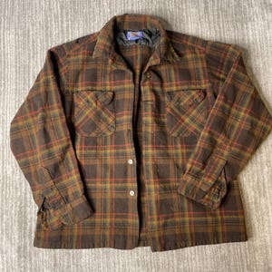 May include: A long-sleeve button-up shirt featuring a brown, orange, and green plaid pattern. The shirt includes a pointed collar, two chest pockets with buttoned flaps, and a Pendleton brand label on the inside neck. The front closure has buttons.