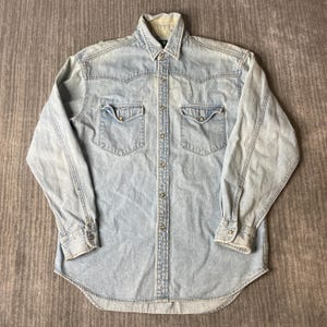 Könnte beinhalten: Ein langärmeliges Hemd aus hellblauem Denim mit spitzem Kragen, einer durchgehenden Knopfleiste mit metallischen Druckknöpfen, zwei Brusttaschen mit geknöpften Klappen sowie Schulterpassen im Western-Stil und geknöpften Ärmelbündchen.