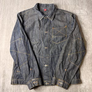 May include: A dark blue denim jacket featuring a classic collar, a front button closure, and long sleeves. The garment is detailed with contrasting yellow stitching, including an X-shaped pattern on the chest pocket and on both elbow areas.