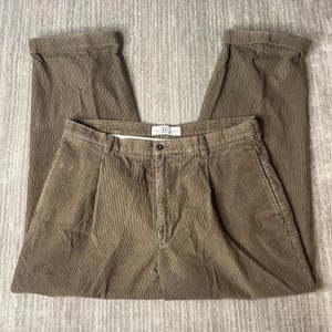 Puede incluir: Pantalones de pana marrón con frente plisado, cierre de botón y cremallera, y dobladillos con vuelta. La cintura incluye una etiqueta interior blanca con el texto EQ GEAR. Estos pantalones tienen bolsillos laterales y trabillas.