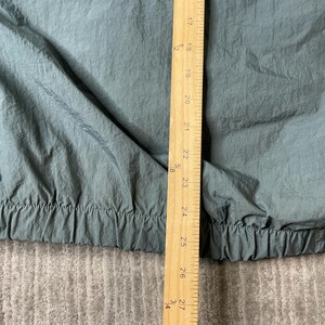 May include: A teal-colored garment with a crinkled texture and an elasticized hem. A wooden ruler is placed vertically over the fabric, showing measurements from 17 inches to 27.25 inches, indicating the length of the item's lower section.