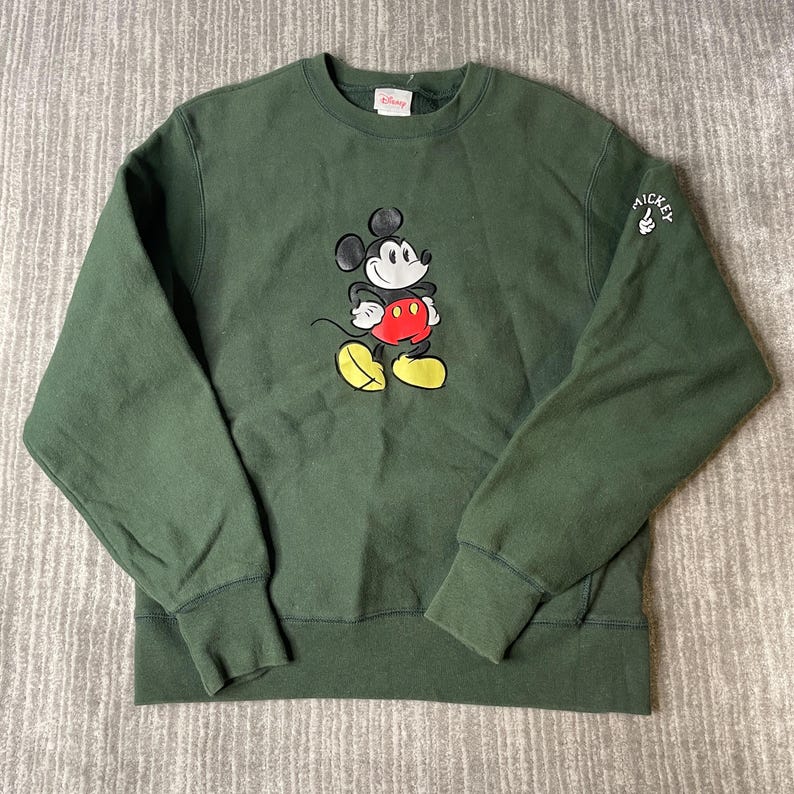 May include: A dark green sweatshirt with a Mickey Mouse graphic. The cartoon mouse has a red belly, yellow shoes, and black ears. The sleeve has the word "Mickey" with a thumbs up. The sweatshirt has a crew neck and ribbed cuffs.