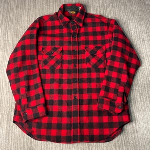 May include: A red and black plaid flannel shirt with two chest pockets and a button-down front. The shirt has a collar and buttoned cuffs. The fabric appears to be a thick, warm material, suitable for cooler weather. The shirt is laid flat on a neutral surface.