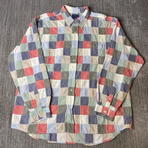 May include: A long-sleeve button-down shirt featuring a multi-coloured checkered pattern in shades of red, blue, green, and cream. The shirt includes a standard collar, a single chest pocket on the left side, and buttoned cuffs on the sleeves.