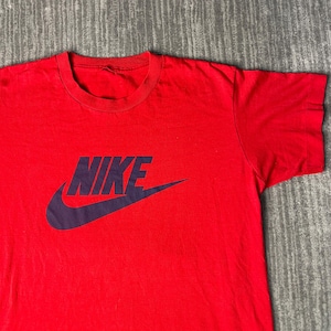 May include: A red t-shirt with a crew neck and short sleeves. The shirt features the word "NIKE" in dark blue block letters with the Nike swoosh logo below the text. The shirt is made of a soft material.