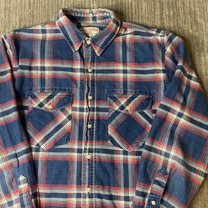 May include: A blue, red, and white plaid button-down shirt with two chest pockets. The shirt has a collared neckline and long sleeves. The fabric appears to be a medium-weight cotton or similar material.