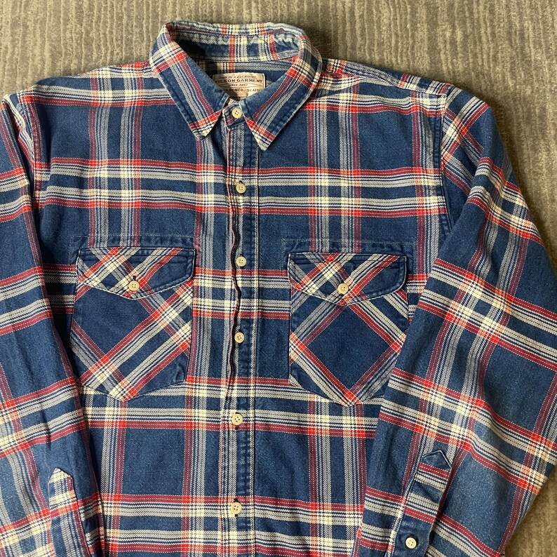 May include: A blue, red, and white plaid button-down shirt with two chest pockets. The shirt has a collared neckline and long sleeves. The fabric appears to be a medium-weight cotton or similar material.