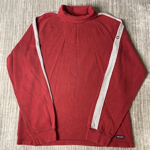 May include: A red long-sleeve mock neck shirt with a ribbed texture. The garment features light grey vertical stripes running down the length of each sleeve. A small circular emblem is visible on the left sleeve, and a rectangular label is at the hem.