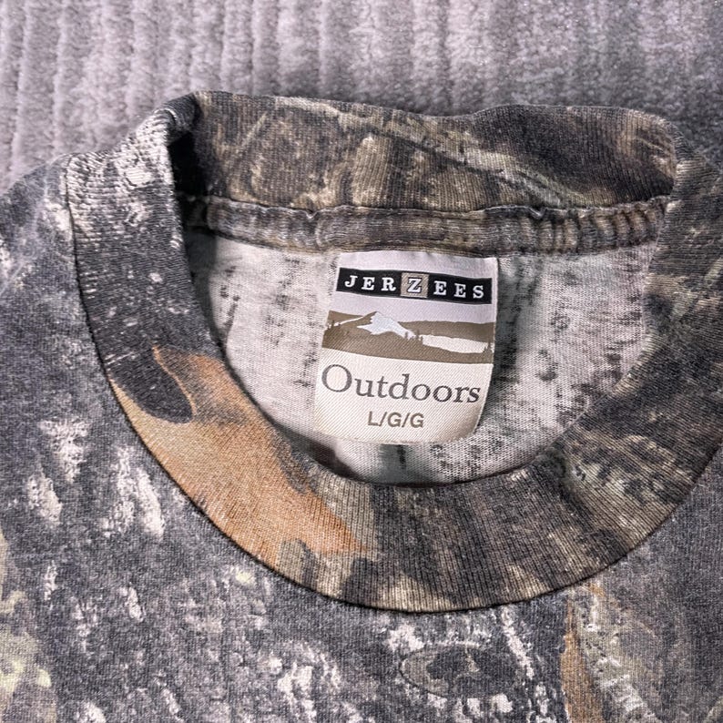 May include: A camouflage-patterned t-shirt with the tag "JERZEES Outdoors L/G/G" visible. The shirt features a blend of brown, gray, and green hues, creating a woodland camouflage design. The collar is visible.