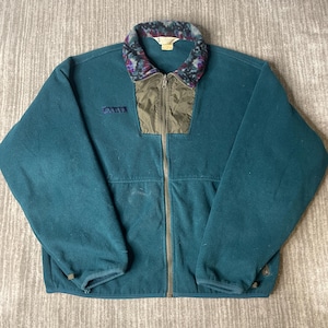 May include: A teal fleece jacket featuring a full-length front zipper, a dark olive green nylon chest panel, and a patterned collar with shades of purple, blue, and black. The jacket includes long sleeves and a Columbia brand logo on the chest.