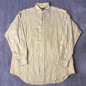 May include: A light yellow button-down shirt with long sleeves. The shirt has a button-down collar and a small blue logo on the chest. The shirt is made of a lightweight fabric.