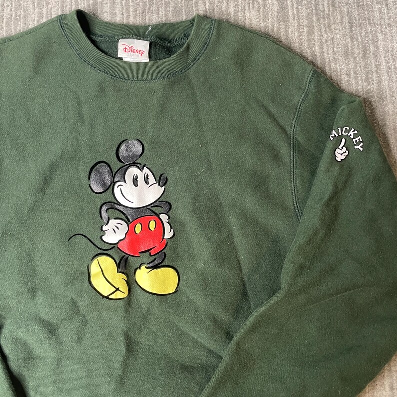May include: A dark green sweatshirt with a Mickey Mouse graphic on the front, wearing red shorts and yellow shoes. The sleeve has a small "Mickey" graphic. The Disney logo is visible on the collar.