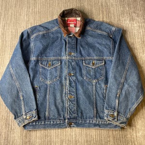 May include: A blue denim jacket featuring a brown leather collar, a red and white plaid interior lining, two chest pockets with button closures, and a front button-down placket. The jacket includes long sleeves with buttoned cuffs.