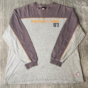 May include: A long-sleeve shirt featuring a grey body and dark grey sleeves with orange piping. The chest displays the text "DCSHOECOUSA" in orange and the number "07" in dark grey. A small DC logo patch is located at the bottom right corner.