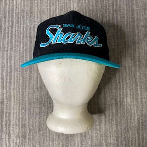 Op de afbeelding: Een zwarte honkbalpet met de tekst SAN JOSE Sharks geborduurd in talingblauw en wit op de voorzijde. De pet heeft een effen zwarte kroon en een platte talingblauwe klep. Het ontwerp bevat de teamnaam in een gestileerd lettertype op de voorkant.