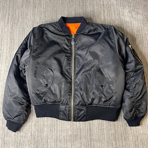 May include: A black bomber jacket featuring a ribbed collar, cuffs, and hem. The jacket has a central brass-toned zipper, two front pockets with snap closures, and a utility pocket on the left sleeve. The interior lining is a bright orange color.