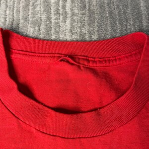 May include: Close-up of a red crew neck t-shirt. The collar and neckline are visible, showing the fabric's texture and stitching details. The shirt is made of a soft, likely cotton material.