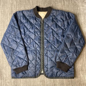 May include: A navy blue quilted jacket featuring a diamond-patterned stitch, a full-length front zipper, and a dark brown ribbed collar and cuffs. The interior lining is a light beige color, visible at the neckline, and the jacket has a straight hem.
