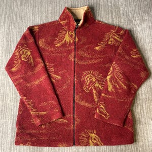 May include: A long-sleeve, full-zip fleece jacket in a deep red color featuring a repeating gold-toned pattern of horse heads. The jacket has a high collar with a tan-colored interior lining and a black zipper closure running down the center front.
