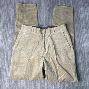 May include: Tan corduroy pants with a classic five-pocket design. The pants are laid flat on a gray surface, showcasing the vertical ribbing of the corduroy fabric. The pants have a button and zipper closure.