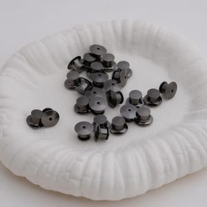 May include: A collection of dark gray metal pin backs, scattered on a white, textured, round surface. The pin backs have a cylindrical shape with a flat, circular base and a post for attaching to pins. The background is a soft, white fabric.