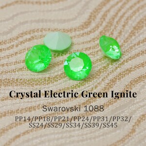 Puede incluir: Primer plano de varios cristales de estrás verde eléctrico facetados. Los cristales tienen varias formas, incluyendo redondas y ovaladas, y están dispersos sobre una superficie texturizada de color beige. El texto de la imagen dice "Crystal Electric Green Ignite" y "Swarovski 1088".