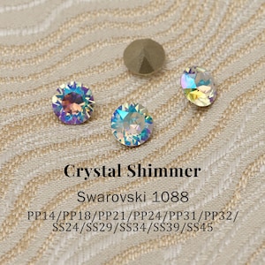 Może przedstawiać: Zbliżenie kilku opalizujących kryształowych dżetów i pojedynczego, matowego, metalicznego ćwieka. Widoczny jest tekst "Crystal Shimmer" i "Swarovski 1088" wraz z kodami rozmiarów.