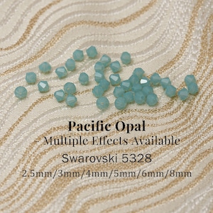 Puede incluir: Una colección de cristales Swarovski Pacific Opal en varios tamaños, que van desde 2,5 mm hasta 8 mm. Las cuentas son de color azul verdoso claro y están dispersas sobre una superficie beige texturizada. El texto de la imagen dice "Pacific Opal - Multiple Effects Available Swarovski 5328".
