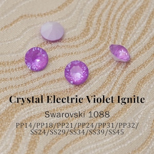 May include: Close-up of several Crystal Electric Violet Ignite Swarovski crystals. The crystals are various sizes and shapes, with a faceted design that reflects light. The text "Crystal Electric Violet Ignite" and "Swarovski 1088" is visible.