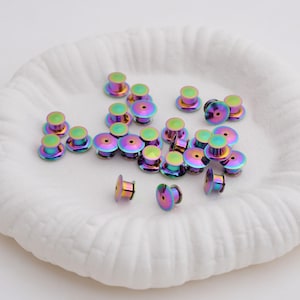 May include: A collection of iridescent pin backs, displaying a spectrum of colors including purple, green, and gold. The pin backs are arranged on a white, textured surface, showcasing their metallic sheen and circular design. These are ideal for crafting and jewelry making.