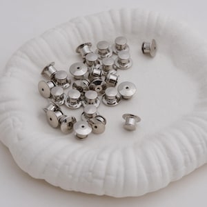 May include: A collection of silver-colored pin backs, also known as clutch backs, displayed on a white, textured surface. The pin backs have a flat, circular base and a cylindrical post for securing pins. The image is well-lit, highlighting the metallic sheen of the objects.