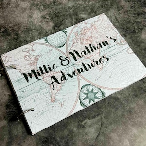Pode incluir: Um diário de viagem personalizado com um design de mapa-múndi vintage. A capa apresenta o texto "Millie & Nathan's Adventures" em escrita elegante. O diário é encadernado com anéis de metal e tem um esquema de cores azul claro e castanho.