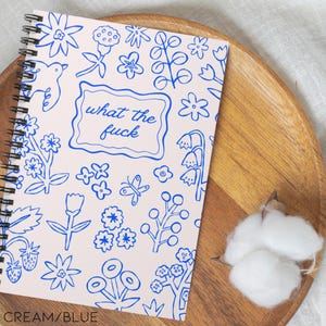 May include: Cream and blue spiral-bound notebook with a floral design. The cover features the phrase "what the fuck" in a blue frame. The notebook is decorated with blue floral and botanical illustrations.