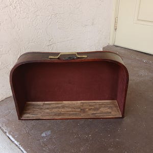 May include: A vintage-style display case with a curved top and a rich, dark brown exterior. The interior is lined with a deep red fabric, and the base features a miniature wooden plank floor. A brass-colored handle adds a touch of elegance.