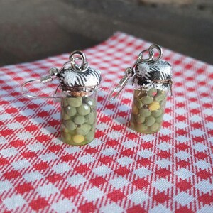 May include: A pair of novelty earrings featuring miniature glass jars filled with green beads and a few yellow beads, resembling olives. Each jar has a cork stopper topped with a black and white checkered fabric. The earrings hang from silver-colored ear wires.