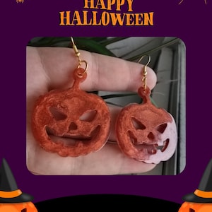 May include: Orange pumpkin-shaped earrings with a carved jack-o'-lantern face. The earrings are made of a translucent material and hang from gold-colored hooks. The background includes the text "HAPPY HALLOWEEN" in orange.