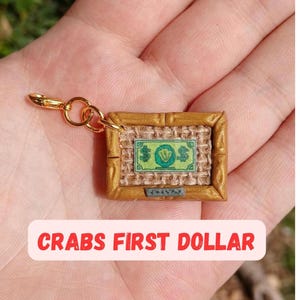 May include: A gold-colored rectangular charm featuring a textured tan burlap background inside a frame. Centered on the burlap is a small green rectangular graphic resembling a dollar bill with a crab icon. A gold lobster clasp is attached.